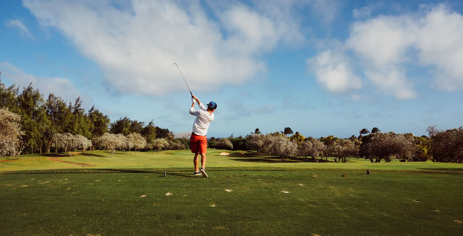 A golfer seen from behind at the end of his drive on a tee, with a wide cloudy sky over the fairway.